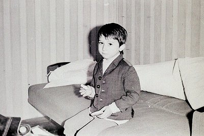 Young boy in mid-20th-century formalwear (vest, button-down shirt) seated on a tufted sofa, holding a small object. Plain str...