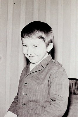 Young boy in a mid-century suit, likely 1950s–1960s. Clean, cropped hair and a relaxed smile against a striped wall backdrop....