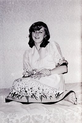 Portrait of a woman in a mid-century formal pose, seated on a patterned floor with floral skirt and lace blouse. Soft lightin...