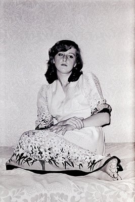 Portrait of a woman in mid-20th century fashion, seated on floral-patterned fabric. She wears a lace-collared blouse with a f...