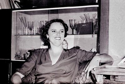 Black-and-white portrait of a woman in a vintage armchair, mid-20th century. She wears a fitted blouse with a high collar and...