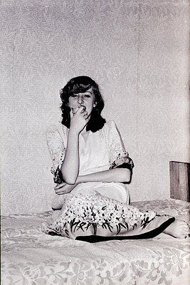 Portrait of a woman in mid-20th century attire, seated on floral-patterned upholstery. She wears a lace-collared blouse and a...