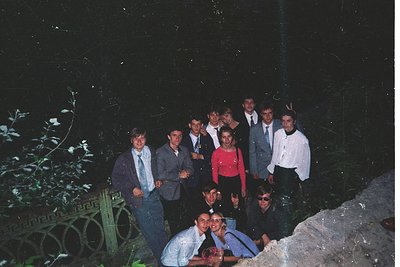 Group portrait in a dimly lit garden setting, likely 1990s. Nine individuals pose outdoors near ornate metalwork and foliage,...