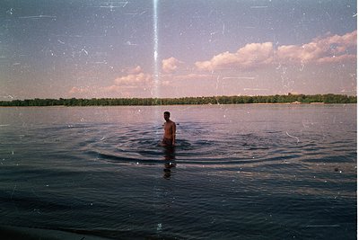 Vintage-style photo of lone figure standing in shallow lake water, surrounded by calm reflections. Distant treeline and open ...