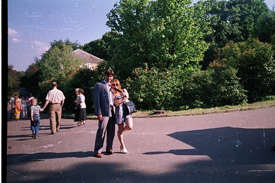 Vintage park scene with mid-20th-century attire. A man in a suit and woman in a knee-length dress pose closely in a shaded pa...