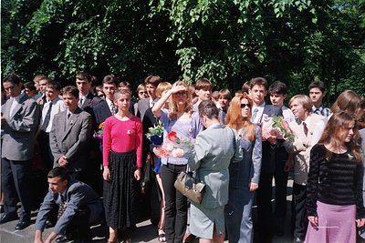 Group graduation ceremony in a wooded park, 1970s Eastern Europe. Uniform attire—men in suits, women in dresses—with floral b...