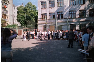 Crowd of men in formal attire (suits, ties) lined up outdoors near a Soviet-era concrete building with barred windows, likely...