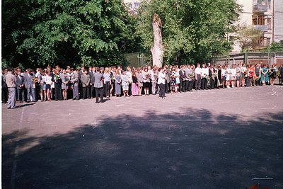 Large outdoor gathering of uniformed individuals, likely students, in a courtyard. Central figure in white dress stands on a ...
