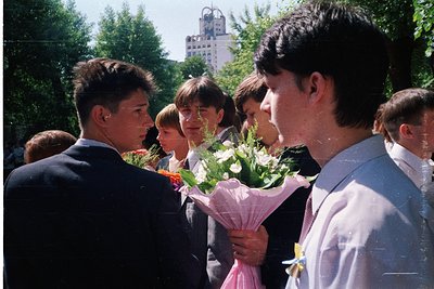 Group of young people in formal attire, likely students, gathered outdoors holding a bouquet of white flowers with green leav...