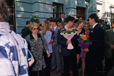 A group of graduates in formal attire poses outside a building, holding bouquets of flowers. Men wear suits with ties, women ...