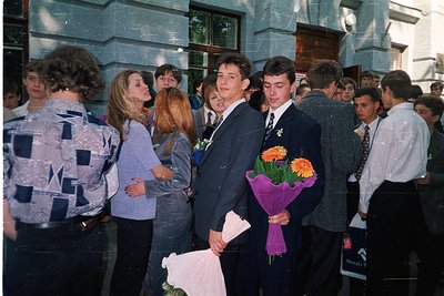 A group of young people in 1980s-1990s formal attire gathers outdoors, likely at a school event. Boys in dark blazers with wh...