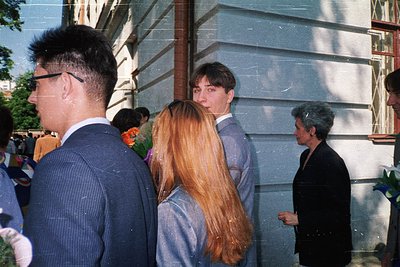 Three individuals in formal attire—men in suits, one with glasses, and a woman in a light blazer—stand outdoors near a concre...