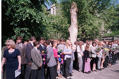 A solemn outdoor gathering in a tree-lined courtyard, likely a graduation ceremony. Students in 1970s-era attire—men in suits...