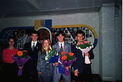 Four graduates pose indoors in formal attire, holding bouquets of flowers. The setting appears to be a school or university w...