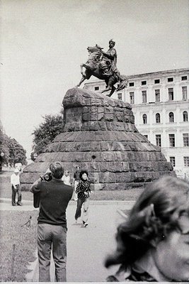 Equestrian statue atop a stone pedestal in a public square, flanked by mid-20th century Soviet-era architecture. Visitors pos...
