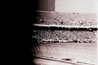 Vintage black-and-white stadium shot featuring Olympic rings logo on track. Mid-20th century stadium with tiered seating, spa...