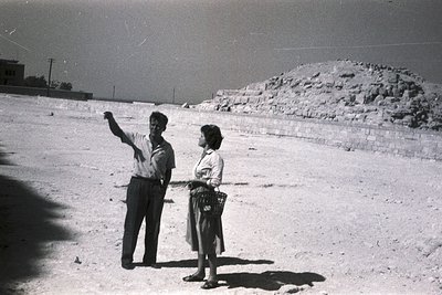 Black-and-white photo of two individuals posing on a sandy, open terrain with a rocky mound in background. Man in short-sleev...