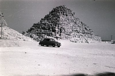 Classic 1950s-era sedan parked beside ancient pyramid ruins in desert terrain. Likely Egypt’s Giza Plateau, showcasing layere...