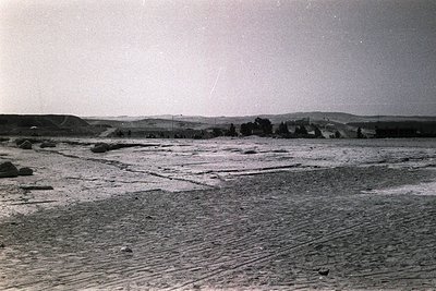 Vintage black-and-white shot of a vast, cracked, dry lakebed or salt flat under clear skies. Distant low-lying hills and spar...