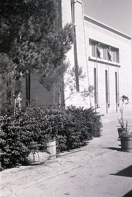 Mid-century modern building facade with Art Deco-inspired vertical columns and large windows. Two figures stand on an upper b...