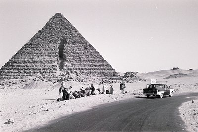 Mid-20th century black-and-white photo of the Great Pyramid of Giza with a vintage car and group of people near the base. Ari...