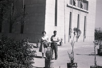 Two individuals pose outdoors near a Brutalist-style concrete building, holding potted plants. The woman wears a skirt and ap...