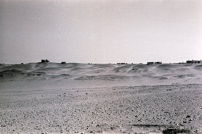 Vast desert landscape with undulating sand dunes and sparse vegetation. A few small, isolated structures—likely traditional o...