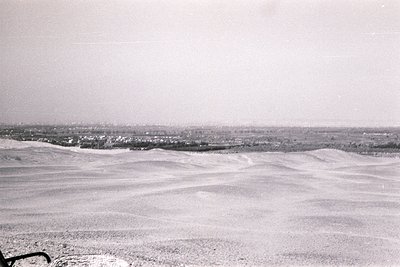 Vast, undulating sand dunes stretch across the foreground, leading to a distant coastal town. The horizon shows faint outline...