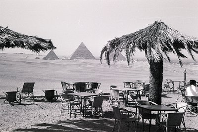 Mid-20th century seaside café overlooking the Great Pyramids of Giza, Egypt. Thatch umbrellas and metal tables/chairs arrange...