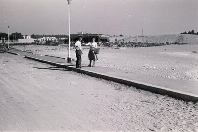 Two individuals in mid-20th-century attire pose near a seaside promenade, likely mid-1950s–1960s. The man wears a suit, the w...