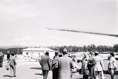 Black-and-white photo of a mid-20th-century outdoor gathering near a small, utilitarian building with a helicopter in the bac...