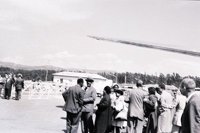 Mid-20th century gathering near a small rural airstrip, featuring a vintage helicopter in background. Group of men and women ...