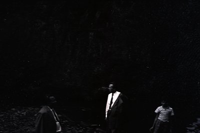 Three silhouetted figures in mid-20th century attire—one in suit, two in casual wear—walking on a gravel path under a starry ...