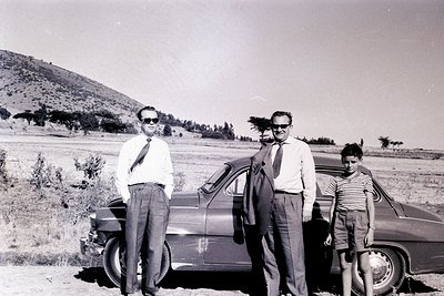 Black-and-white snapshot of three individuals posing beside a vintage sedan in an open, rural landscape. Two men in formal at...