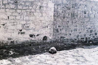 Black-and-white shot of weathered stone wall with rough-hewn blocks, likely 19th–early 20th century. A lone, partially buried...