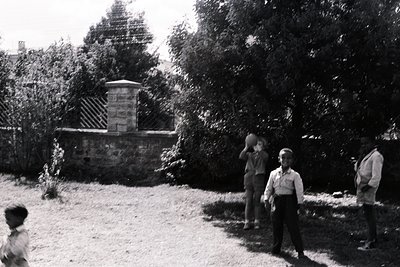 Mid-20th century black-and-white photo: three boys playing near a stone pedestal with striped pattern, surrounded by dense gr...