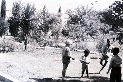 Mid-20th century black-and-white photo of children playing outdoors in a lush, tropical garden. A woman in a light dress stan...