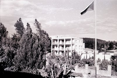 Mid-century concrete apartment block with red-tiled roofs, framed by dense greenery and a flagpole displaying a tricolor flag...