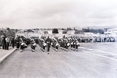 Military band marching in formation, likely mid-20th century. Uniforms feature dark trousers, white shirts, and peaked caps. ...