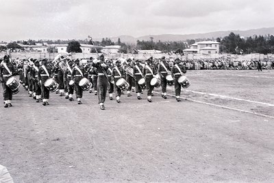 Military band marching in formation, likely 1950s–1960s. Uniforms feature peaked caps, dark trousers, and white belts; drums ...