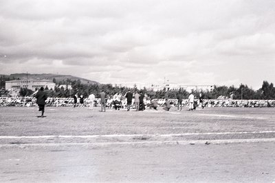 Mid-20th century seaside promenade with stone seawall. A lone figure in dark attire walks left, while groups gather near the ...