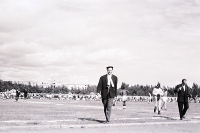 Black-and-white photo of a man in a suit and cap walking across an open field, likely a stadium or sports venue. Crowds gathe...