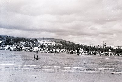 Black-and-white shot of a mid-20th-century stadium event, likely a baseball game. Pitcher in motion on the mound, spectators ...