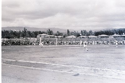 Vintage black-and-white photo of a mid-20th-century soccer match on a dirt track. Three players in shorts and socks, one drib...