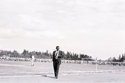 Black-and-white photo of a man in a suit and tie walking on a grass stadium, likely mid-20th century. Crowd of spectators fil...