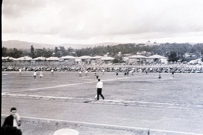 Black-and-white shot of a mid-20th century stadium with athletes in motion on a dirt track, surrounded by temporary bleachers...
