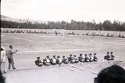 Coach directs youth soccer team in midfield drill, 1960s-70s. Uniforms feature dark jerseys with white trim; spectators fill ...
