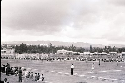 Black-and-white soccer match in a rural stadium, likely 1950s–1960s. Refereeing action on a grass pitch with spectators seate...