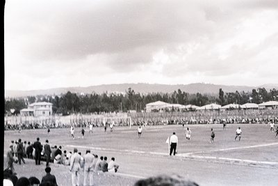 Black-and-white soccer match in a large stadium, likely 1950s–1960s. Players in striped jerseys and shorts engage in action o...