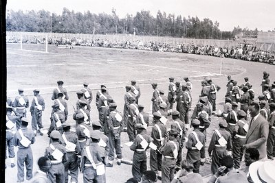 Black-and-white photo of a military formation on a grassy stadium field, likely mid-20th century. Uniformed soldiers in caps ...